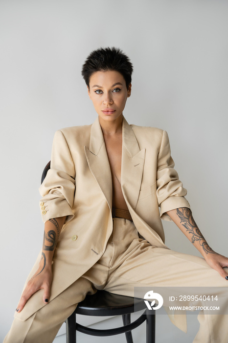 trendy brunette woman in beige pantsuit sitting on chair and looking at camera on grey background.