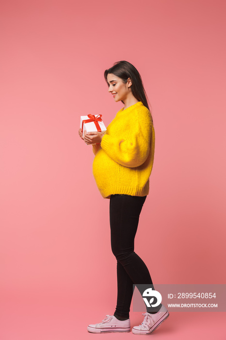 Cheerful young pregnant emotional woman posing isolated over pink background holding gift box.