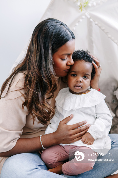 Happy young Indian mother playing kissing mixed race black baby girl daughter. Family mixed race people mom and kid together hugging and embracing at home. Ethnic diversity family.