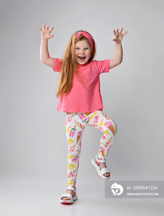 Playful, frolic redhead kid girl in pink t-shirt, colorful pants and sandals plays dino, monster roaring, holding hands with claws up and moving, walking straight towards camera