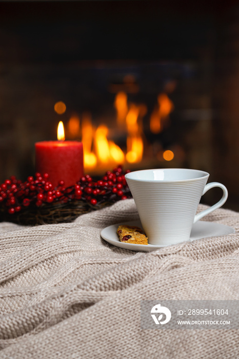 Cup of hot drink with cookie berries and red candle in red Christmas decoration on cozy knitted plaid in front of fireplace. Christmas New Year concept. Cozy relaxed magical atmosphere home interior.