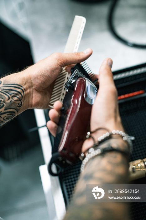 Cropped view of tattooed barber holding comb and hair clipper in barbershop