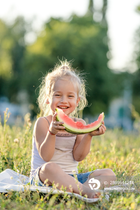 Little blond girl eating watermelon in the park.