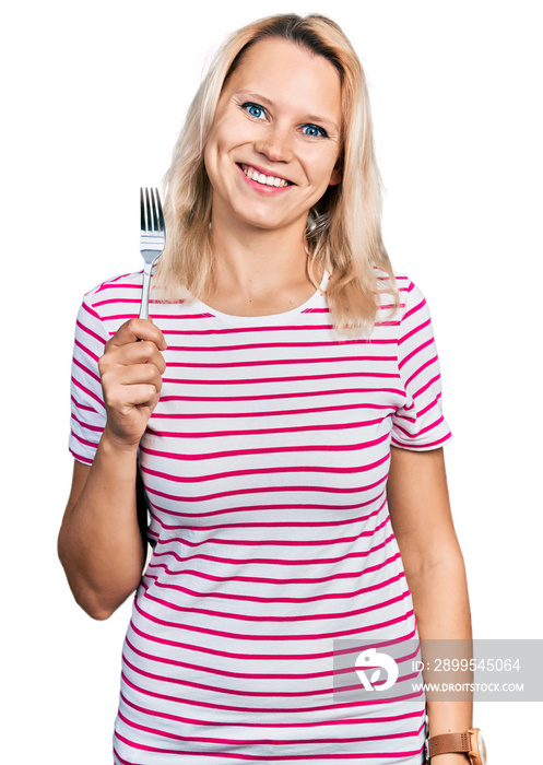 Young caucasian woman holding one silver fork looking positive and happy standing and smiling with a confident smile showing teeth