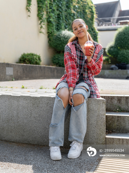 Portrait of young woman with smoothie sitting on stone wall