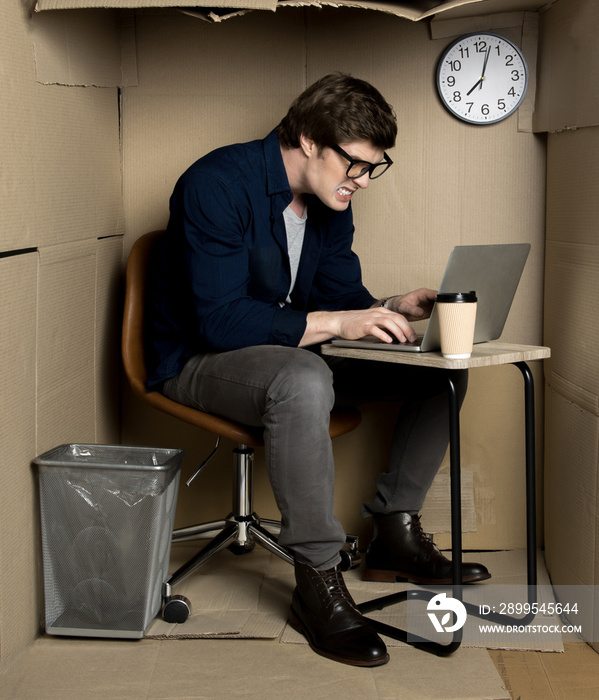 Feeling furious. Full length of young employee is expressing anger while typing on modern laptop. He is sitting at workplace in cramped cardboard office. Business trouble concept