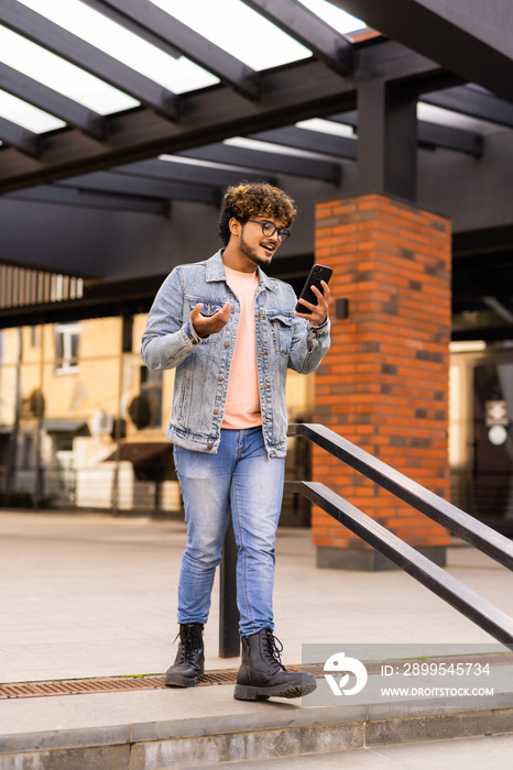 Indian man using mobile phone waving hand, communication online on the street. Happy successful asian influencer recording video outdoors