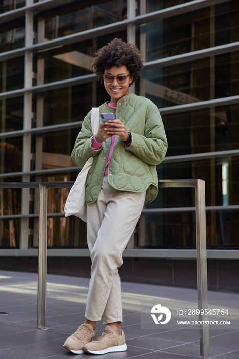 Fashionable positive female blogger holds mobile phone uses modern technology for phoning online and networking social media wears trendy sunglasses stylish outfit stands outside sends sms to friend