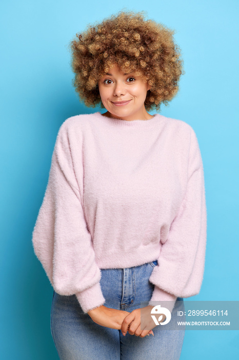 Vertical shot of gentle young woman with curly hair keeps arms together looks shy wears casual fluffy sweater and jeans isolated over blue wall