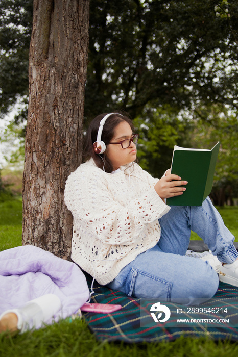 Young woman with Down syndrome reading and listening to music in the park