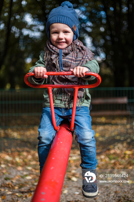 Little cheerful boy in a blue cap on a red seesaw