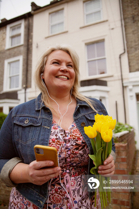 Happy plus-sized woman holding flowers while video-chatting on her phone in a neighborhood