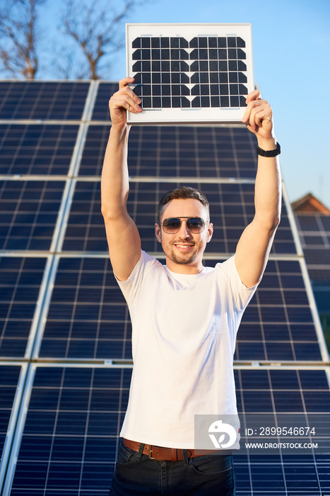 Smiling young man holding a small pv module above his head looking at the camera, solar plant behind his back, concept of alternative energy