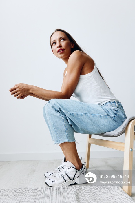 A woman sitting on a full-length chair in a studio against a white wall with very tanned skin in blue jeans and a white T-shirt