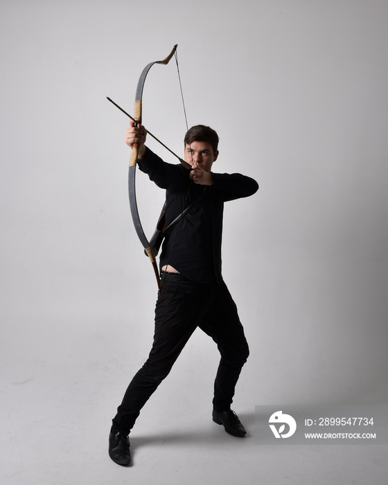 Full length portrait of a  brunette man wearing black shirt and waistcoat holding  a bow and arrow.  Standing  action pose isolated  against a grey studio background.