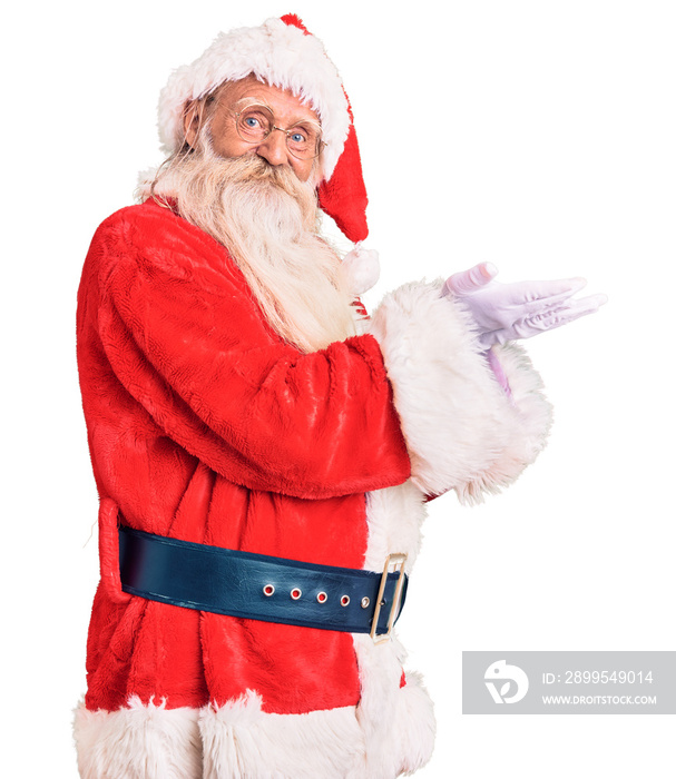 Old senior man with grey hair and long beard wearing traditional santa claus costume pointing aside with hands open palms showing copy space, presenting advertisement smiling excited happy