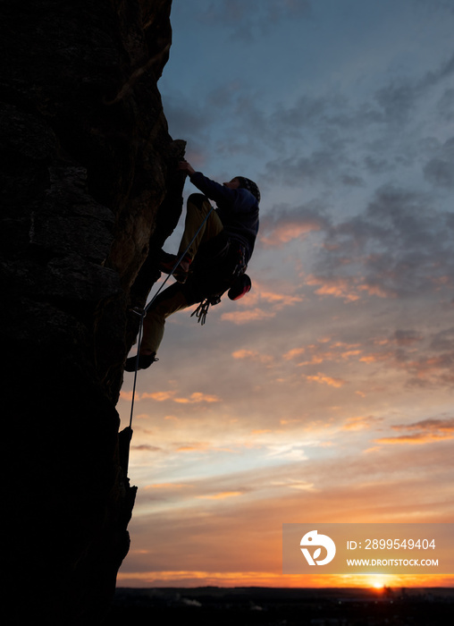 Male climbing in safety harness, looking up and taking decisive step on challenging rocky route. Side view. Wonderful sky during sunset on background. Copy space. Rock climbing and leadership concept