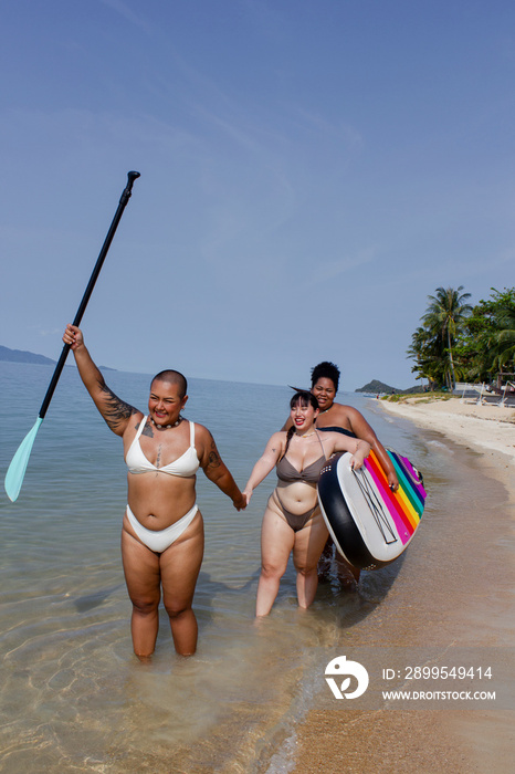 Group of cheerful women walking with paddleboard along beach