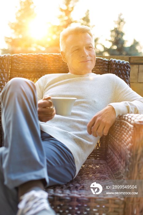 Smiling mature man holding drink while sitting on wicker chair at patio