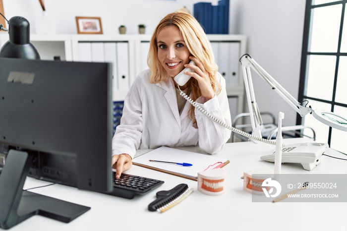 Young blonde woman wearing dentist uniform speaking on the phone at dental clinic