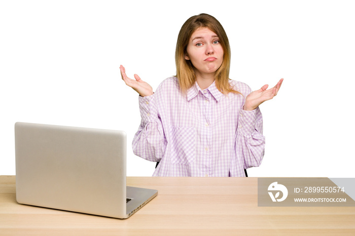 Young caucasian woman in a workplace working with a laptop isolated doubting and shrugging shoulders in questioning gesture.