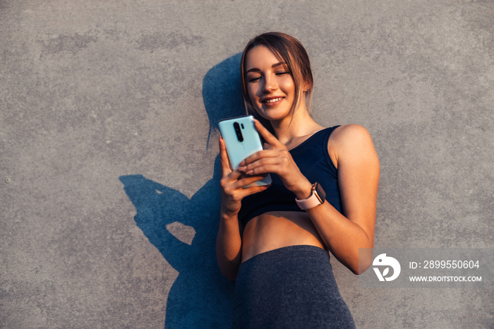 Smiling fitness woman, with smart watch uses mobile phone, looks at smartphone against gray city wall background