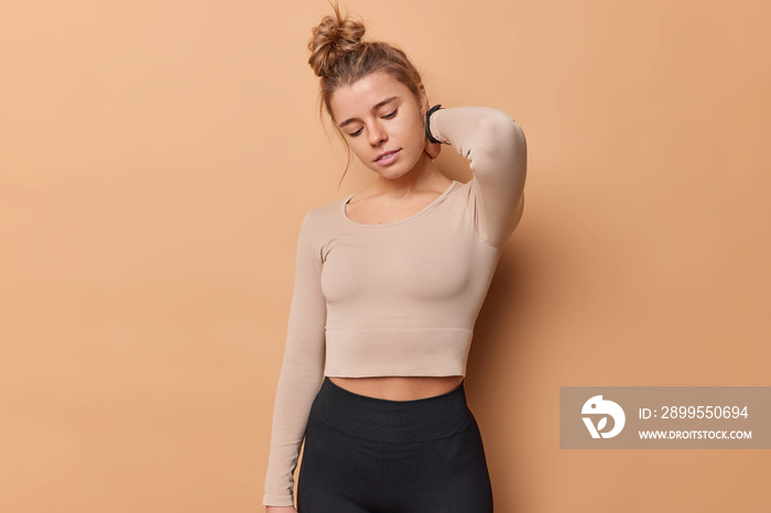 Sporty woman tired after fitness training keeps hand on neck feels pain wears cropped top and leggings focused down isolated over beige studio wall being in good physical shape. Workout indoor