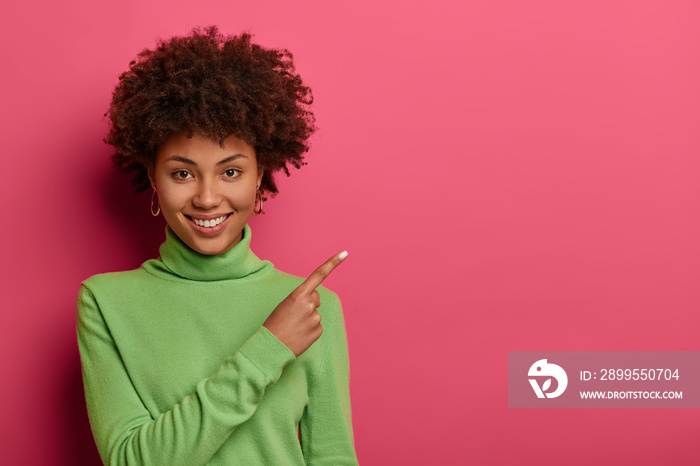 Adorable woman with curly hairstyle, healthy skin and white teeth, points on blank place, shows space for your advertising content, wears casual green sweater, gives direction or way on right