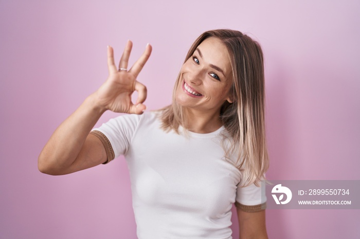 Blonde caucasian woman standing over pink background smiling positive doing ok sign with hand and fingers. successful expression.