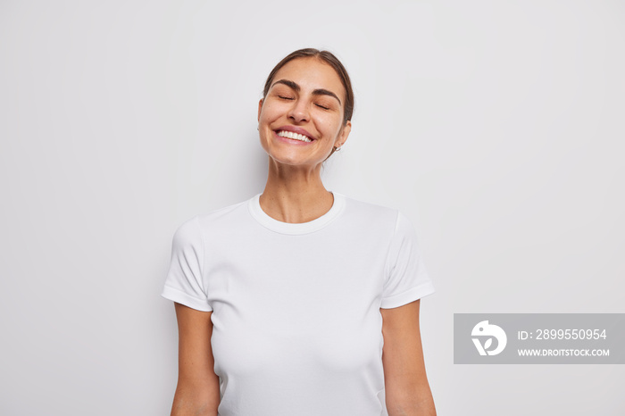 Sincere positive European woman with dark hair keeps eyes closed smiles broadly shows white teeth imagines something good dressed in casual t shirt poses indoor. Happy human emotions and feelings