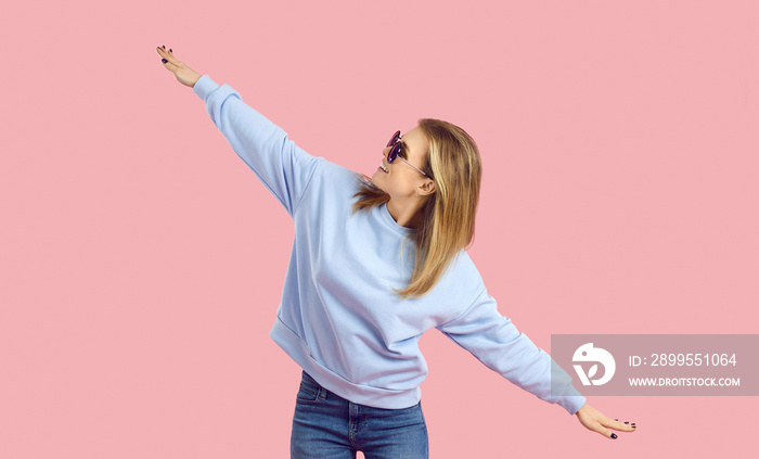 Enjoying music and catching the good vibes. Happy young woman having fun in a fashion studio. Carefree girl in sunglasses and pale blue sweatshirt dancing isolated on a pastel pink background