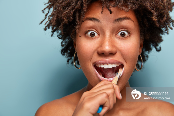 Dental hygiene and dentistry concept. Headshot of surprised young Afro American woman with crisp hair, uses toothbrush and toothpaste for cleaning teeth, stares with bugged eyes isolated on blue