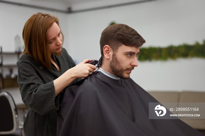 Handsome blue eyed man sitting in barber shop. Hairstylist Hairdresser Woman cutting his hair. Female barber.