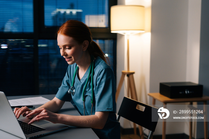 Side view of happy cheerful redhead young female doctor in blue green medical uniform working typing on laptop computer, sitting at desk in dark hospital office room near window at night.