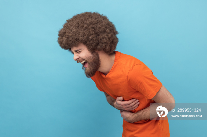 Side view of laughing man with Afro hairstyle in orange T-shirt holding his stomach and hunched in crazy hysterical laughter, sincere joyful emotions. Indoor studio shot isolated on blue background.
