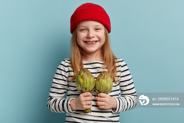 Isolated shot of cheerful lovely girl carries artichoke, helps mother with preparing fresh spring salad, wears stylish red hat and striped jumper isolated over blue background. Vegeterian food concept