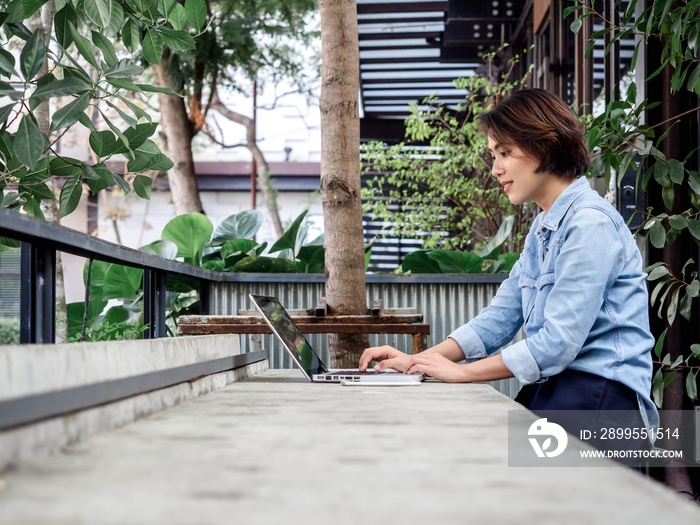 Beautiful happy asian woman short hair wearing blue jeans shirt using laptop computer in cafe with green background.