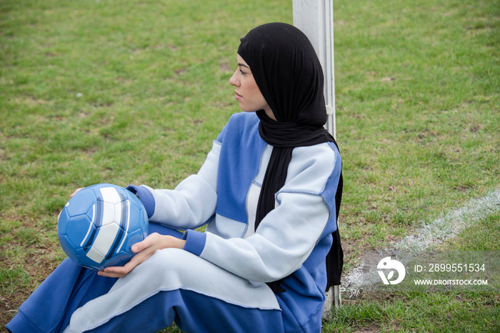 Portrait of woman in hijab sitting in soccer field with soccer ball