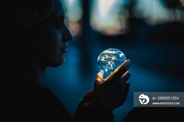 young worried woman holding bright shining lightbulb in her hands feeling unsave while in fear thinking about a future blackout with no electricity or power outage with blue cold background