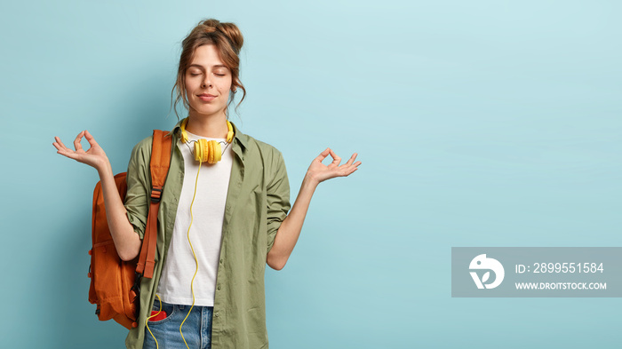 Photo of relaxed student keeps hands in mudra gesture, keeps eyes closed, listens peaceful music, headphones on neck, wears shirt and jeans, has rucksack, isolated on blue background with free space