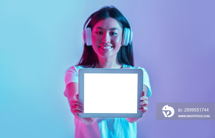 New generation of devices. Smiling asian girl in headphones shows tablet with blank screen on pink and blue background in light of neon