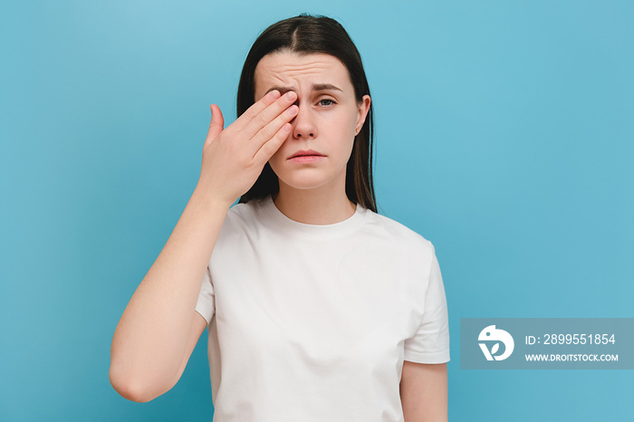 Young girl suffering from eyes pain and feeling something in eye, posing on blue wall. Cause of pain include contact lens problem, conjunctivitis, foreign object, dry eye syndrome or allergy