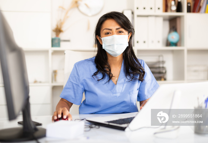 Portrait of female nurse wearing face mask sitting at doctor room and chechking appointments in her laptop