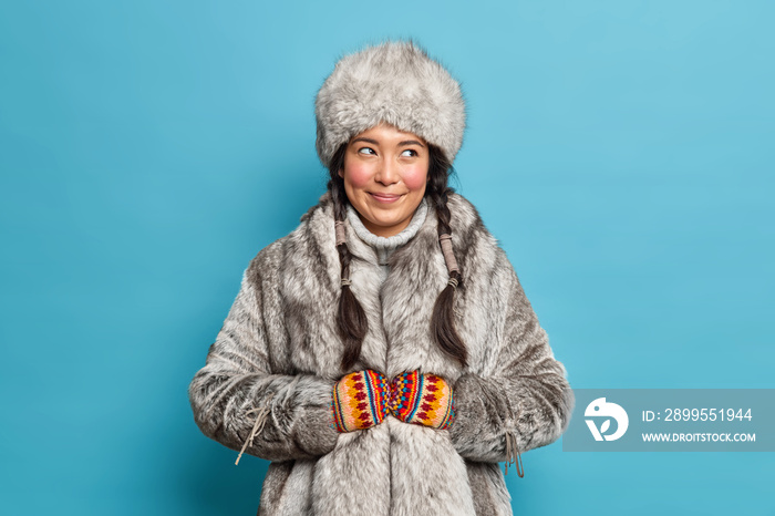 Pleased smiling eskimos woman dressed in traditional clothing adapted to extreme cold lives on nothern pole isolated over blue background. Female inhabitant of Alaska wears fut coat and hat.