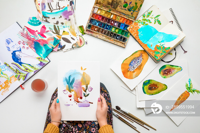 top view of woman holding watercolor drawing while surrounded by color pictures