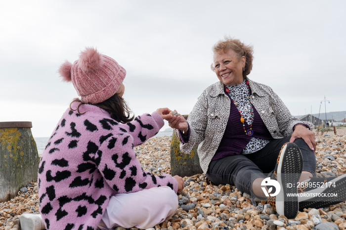 Grandmother giving stone to granddaughter while sitting on beach on cloudy day