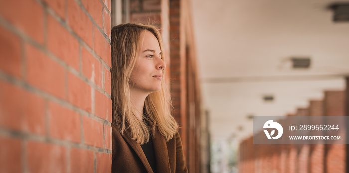 Cute fashionable young woman in a red brick hallway looking away in the distance. Caucasian girl in a let’s go mode, wanting to travel. Wanderlust hipster girl smiling. Copy space on the left.