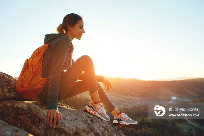 woman hiker with backpack sits on edge of cliff against background of sunrise