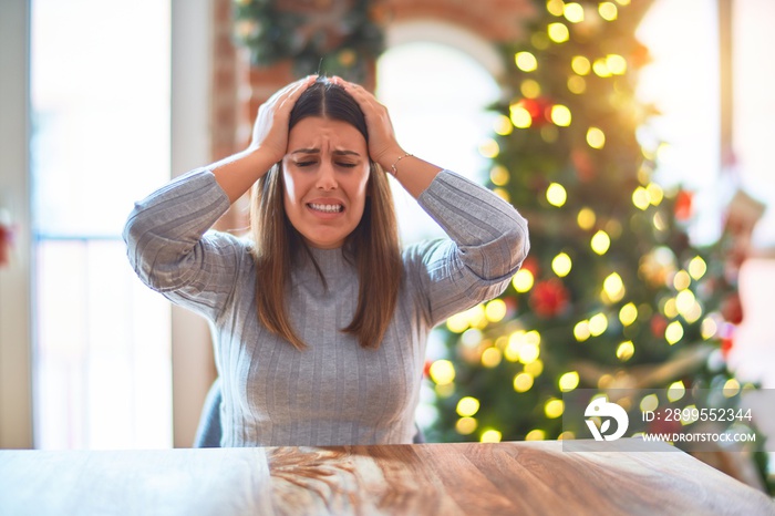 Young beautiful woman sitting at the table at home around christmas tree and decoration suffering from headache desperate and stressed because pain and migraine. Hands on head.