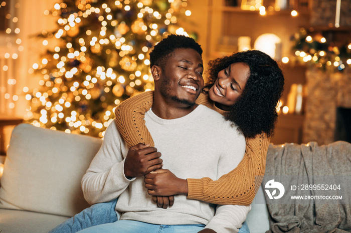 Affectionate young black couple hugging on sofa in decorated room with magic lights on Christmas